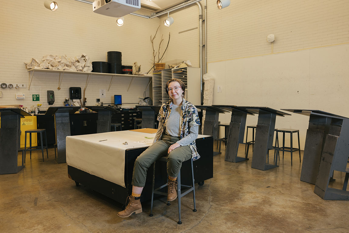 A white person with short black hair, glasses and quirky business casual clothing sits casually in a drawing studio